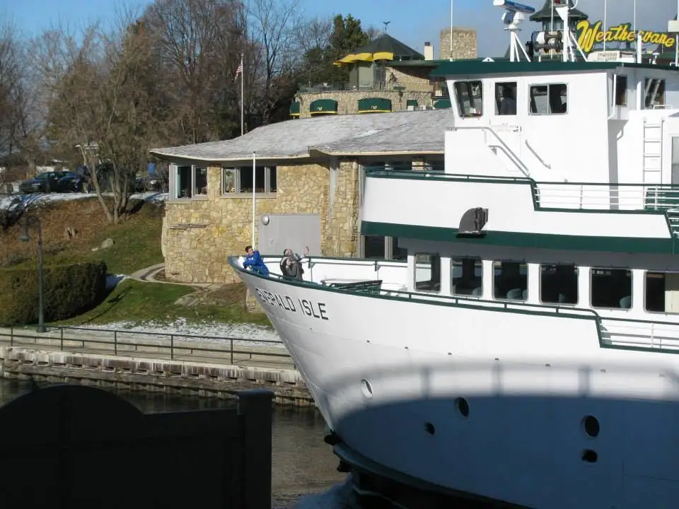 A white ferry beside a stone waterfront building, with people on the bow and lettering on the vessel and sign.