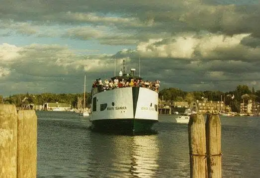 A ferry approaches between wooden pilings with passengers crowded along its upper deck.