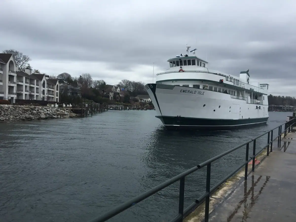 A large white ferry marked “EMERALD ISLE” approaches a wet waterfront pier beneath an overcast sky.