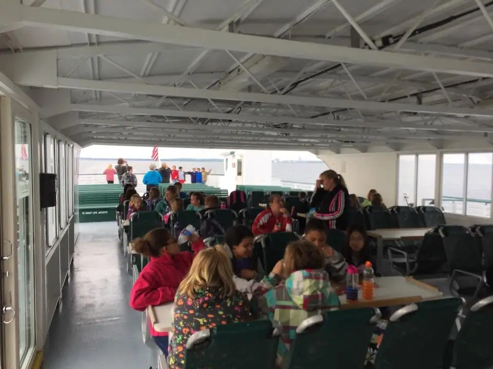 Children and adults sit at tables inside a passenger ferry, with water and American flags visible through the windows.
