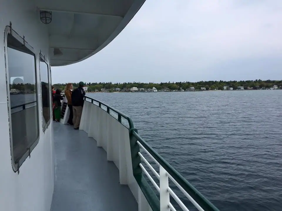 Passengers stand on the open deck of a ferry beside rippling water, facing a wooded shoreline with houses.