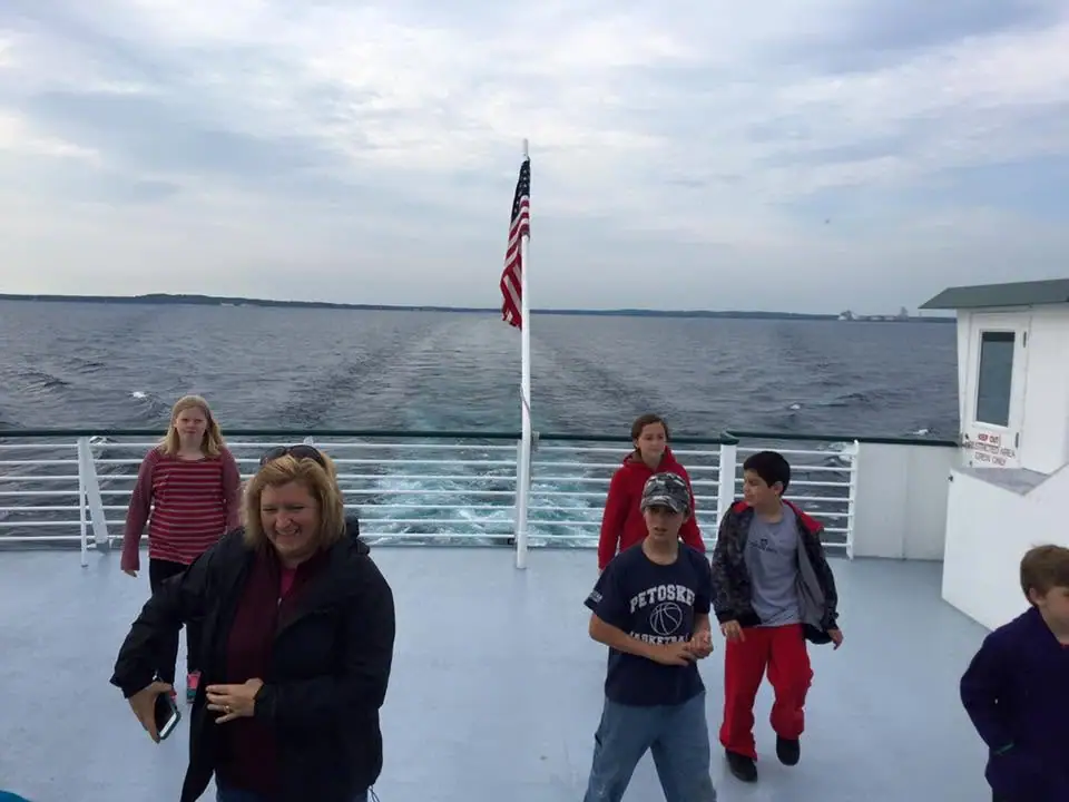 Passengers and children on a ferry deck beneath an American flag, with a wake and distant shoreline behind them.
