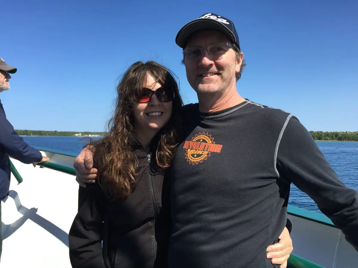 Two adults pose on a boat deck beside the water, with a wooded shoreline behind them.
