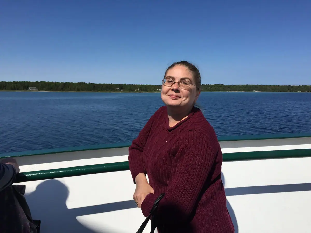 A smiling woman in a burgundy sweater stands beside a boat railing with wooded shoreline behind her.