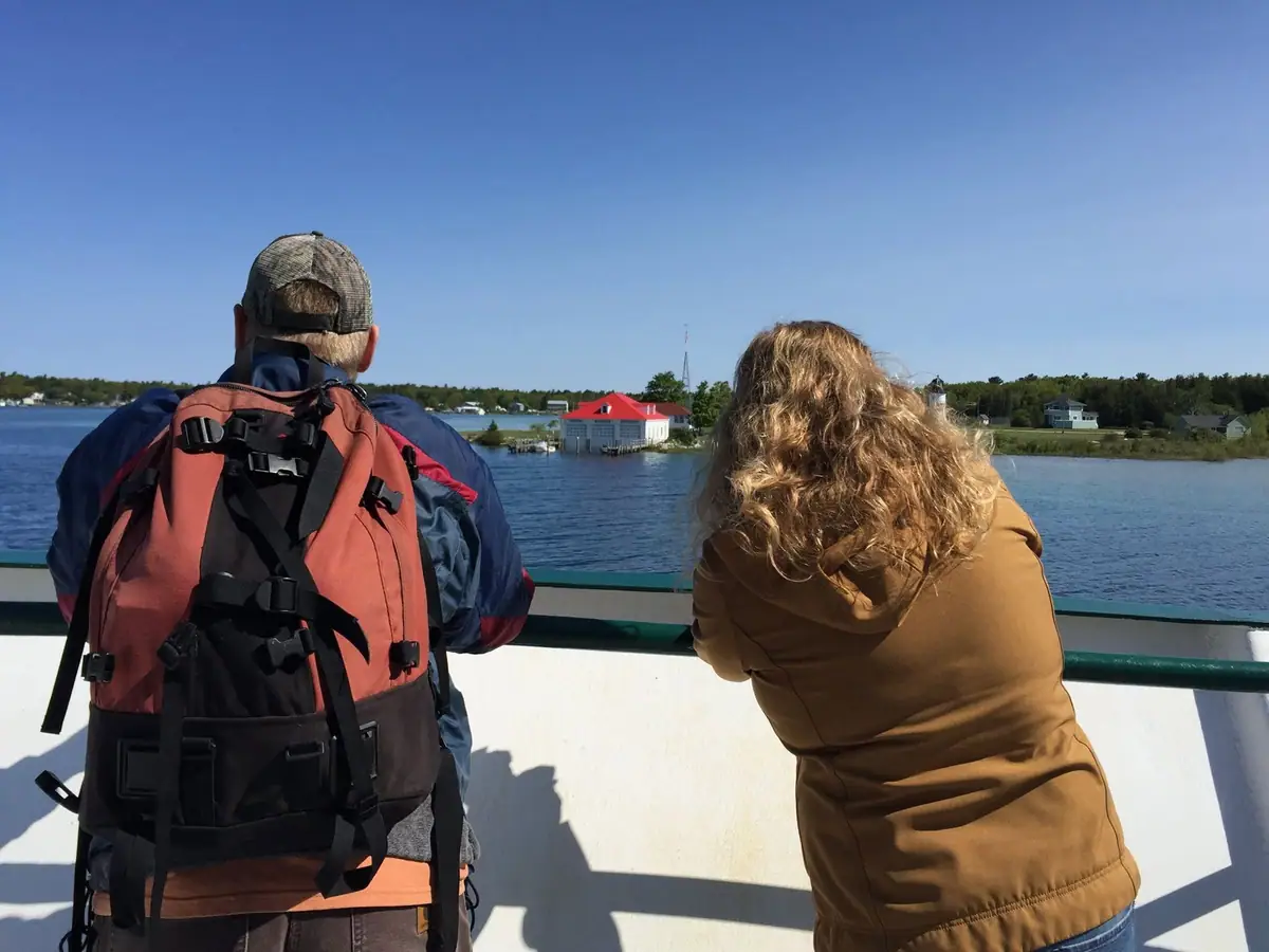 Two passengers stand at a vessel railing, looking toward a red-roofed waterfront building and wooded shore.