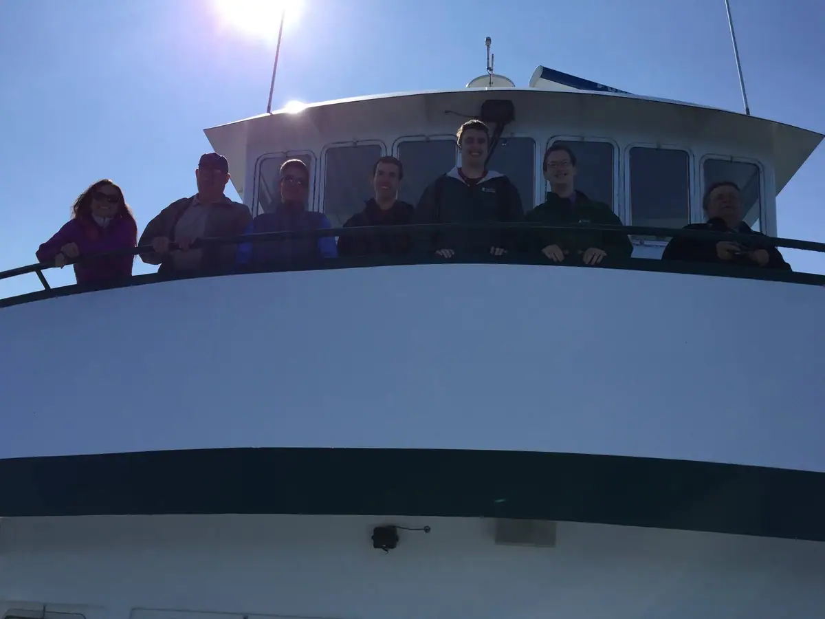 Seven people pose behind a railing above the white-and-dark hull of a vessel in bright sunlight.