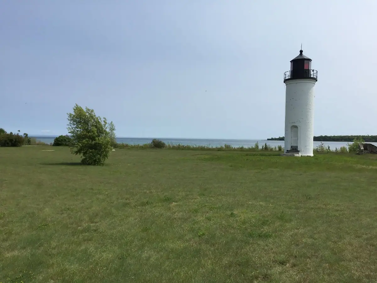 A white cylindrical lighthouse stands in a grassy field beside a broad body of water.