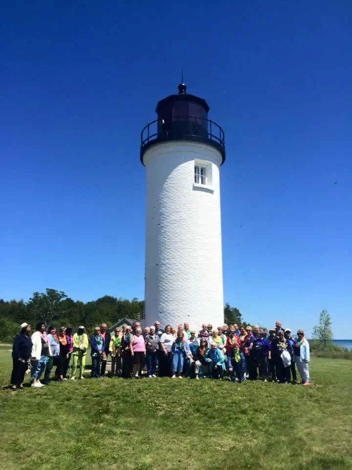 A large group poses on a grassy lawn in front of a tall white lighthouse under a clear blue sky.
