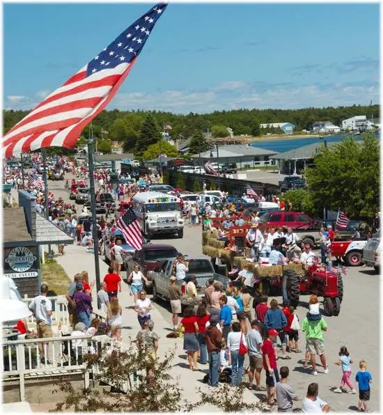 A crowded patriotic parade passes beneath a huge American flag beside a waterfront.