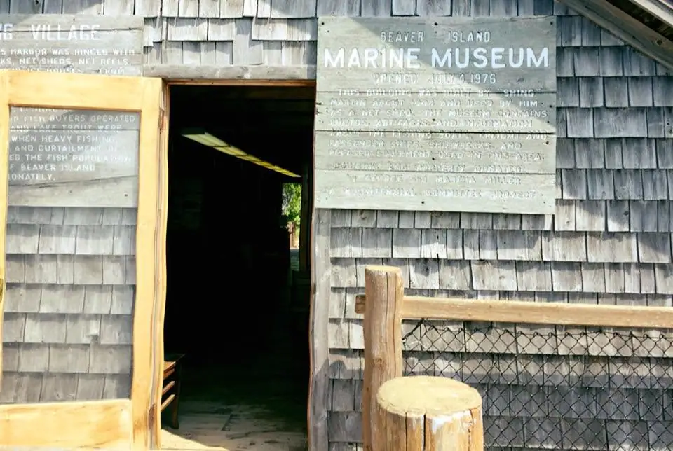 A shingled wooden building with an open doorway, a chain-link fence, and weathered museum signs.