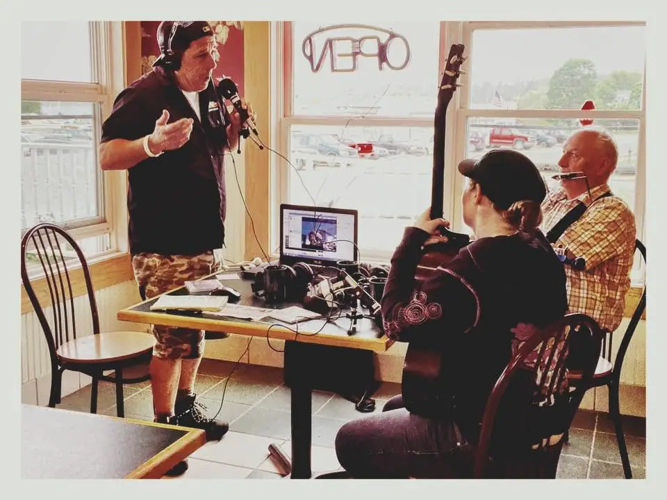 Three people conduct an indoor radio broadcast around a table covered with microphones and audio equipment.