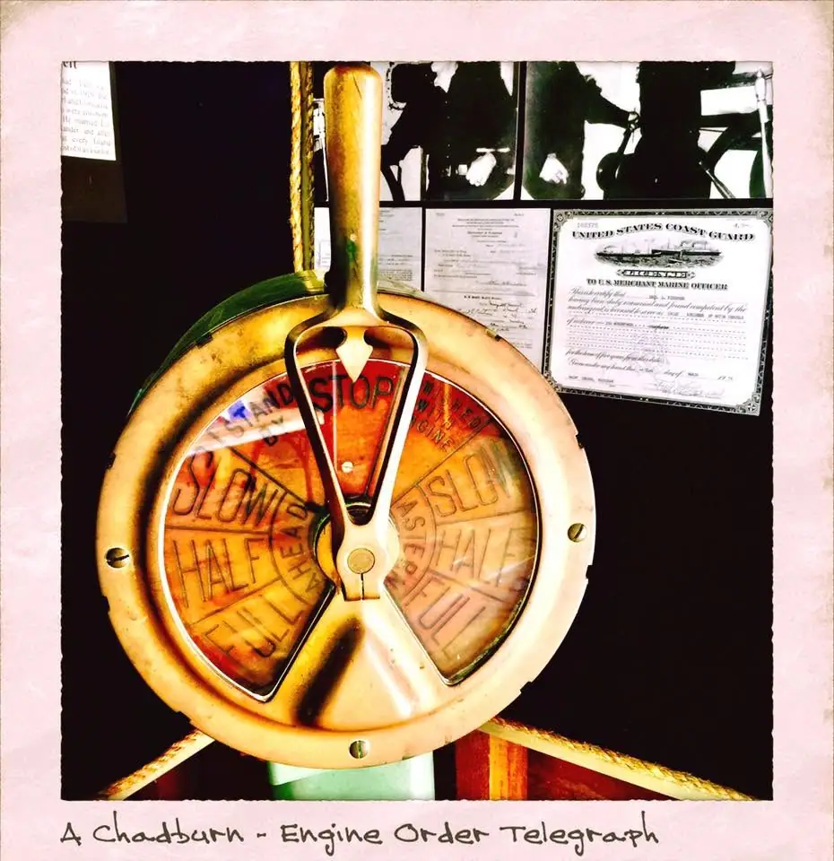 A brass engine-order telegraph with speed markings is displayed beside framed maritime certificates.
