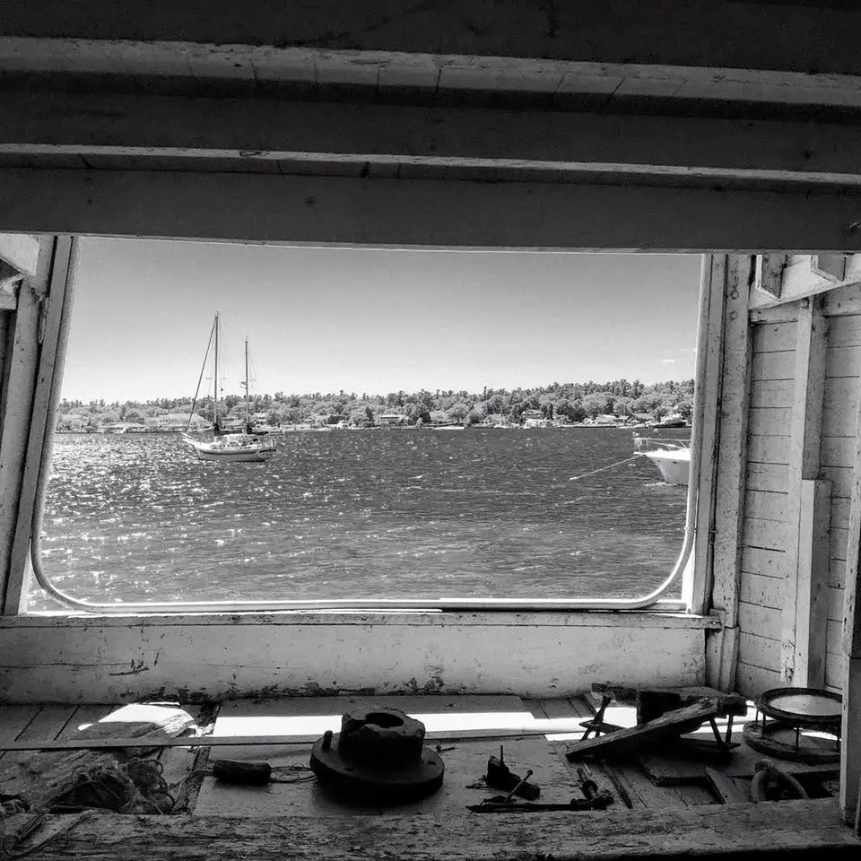 View from a weathered boat interior through a large window toward sailboats, water, and a wooded shoreline.