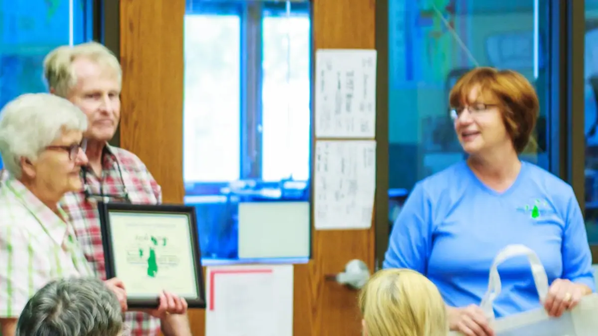 Three adults gather indoors as a man holds a framed certificate beside a smiling woman in a blue shirt.