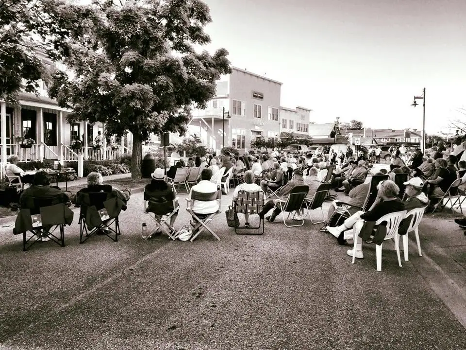 A large seated crowd gathers outdoors on a street beside a porch and downtown buildings.