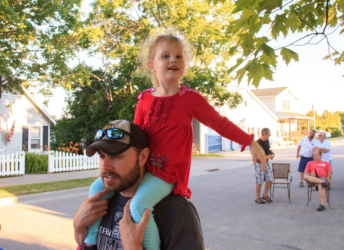 A man carries a young girl on his shoulders while adults gather along a tree-lined residential street.