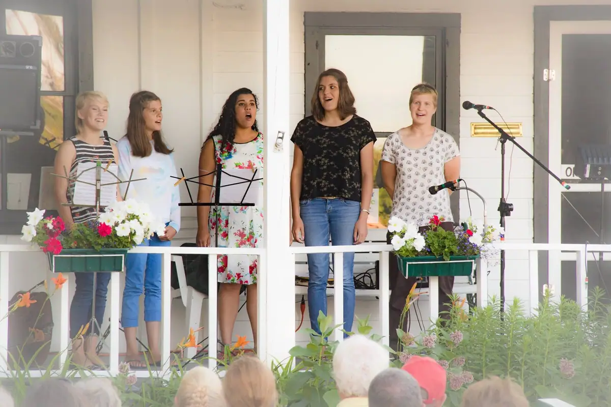 Five women sing on a porch behind music stands and microphones while an audience watches.