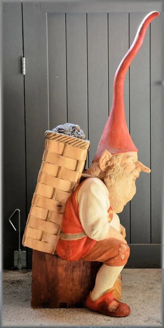 Photograph of a seated bearded gnome sculpture with a tall red hat and a large woven basket.