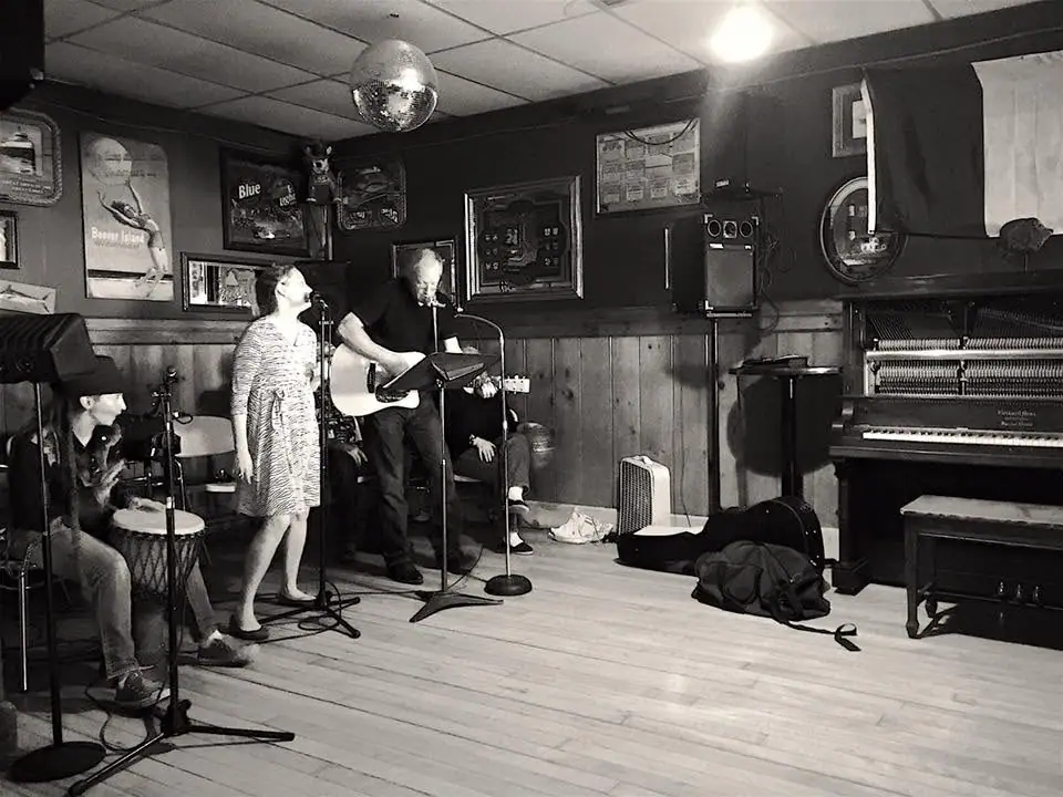 Musicians perform with guitar, hand drum, microphones, and piano in a wood-paneled room.