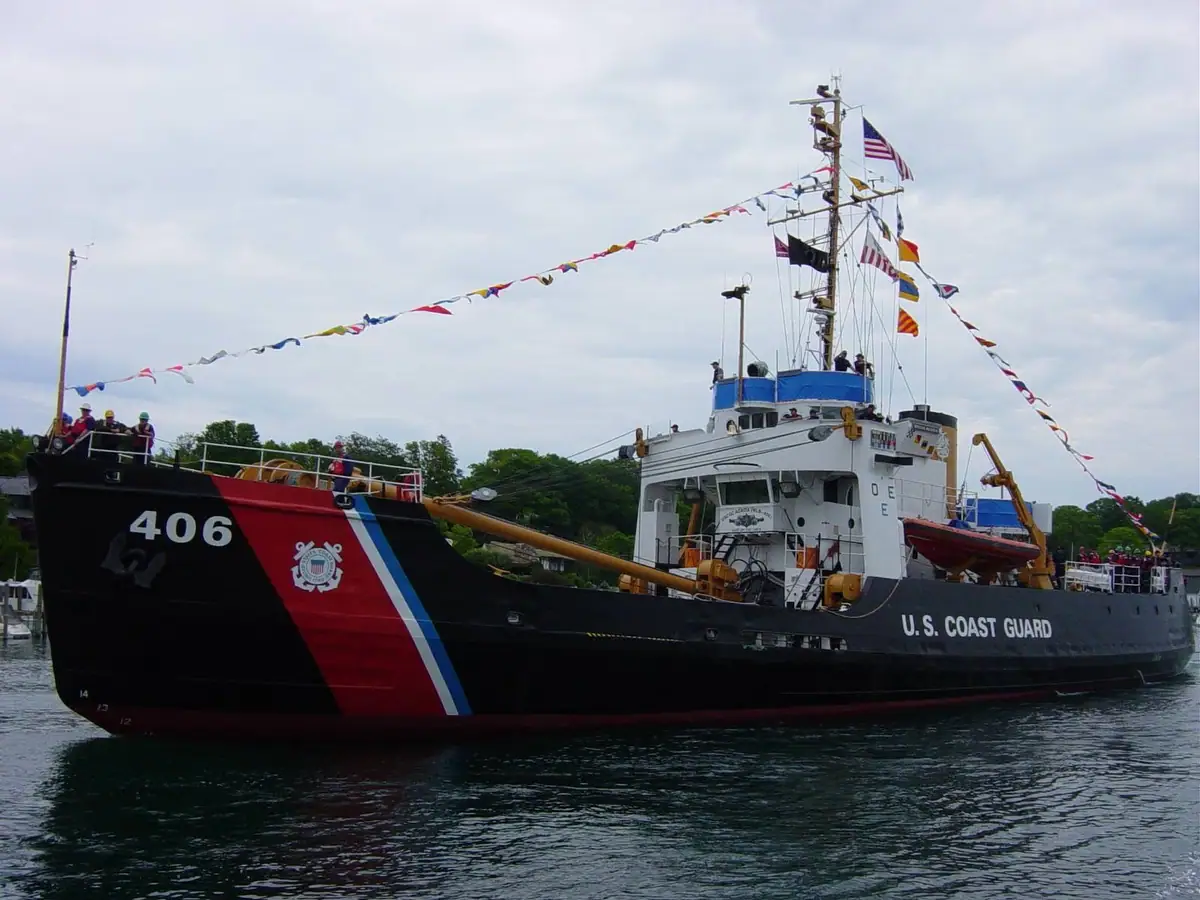 A decorated black-hulled cutter with signal flags and people aboard moves through the water.