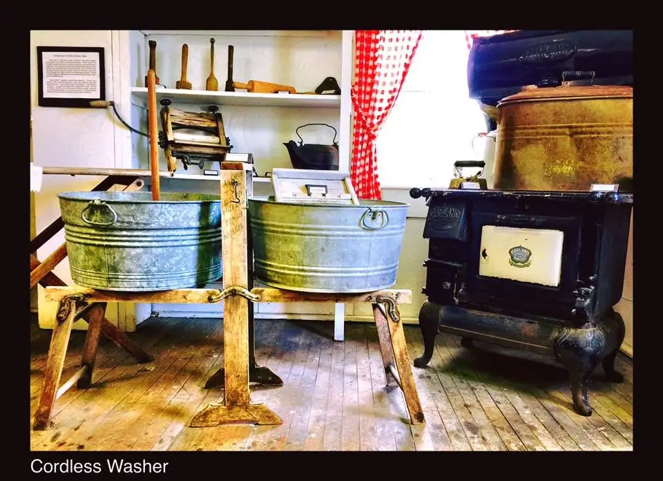 Room display with two galvanized wash tubs, wooden laundry tools, and a cast-iron stove labeled “Cordless Washer.”