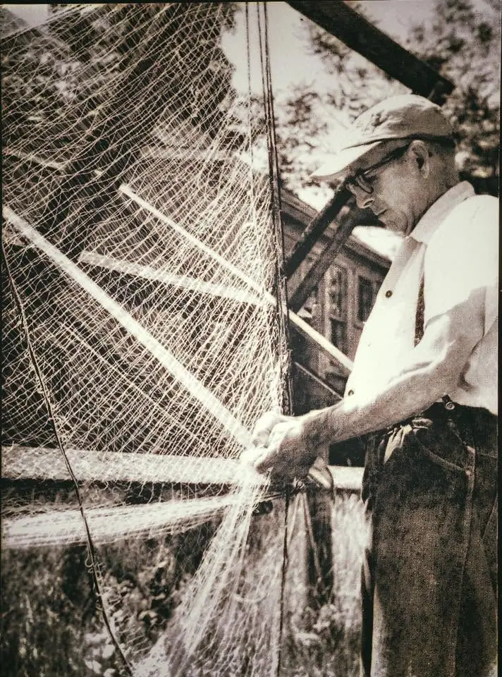 A man wearing a cap and glasses repairs a large fishing net stretched across a wooden frame.
