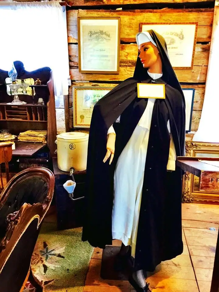 A mannequin in a black-and-white religious habit stands among framed documents and furnishings in a wood-paneled room.