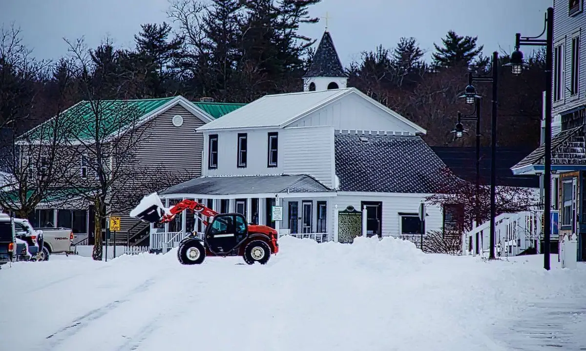 Snow-covered street with a red loader, white church, and surrounding buildings.