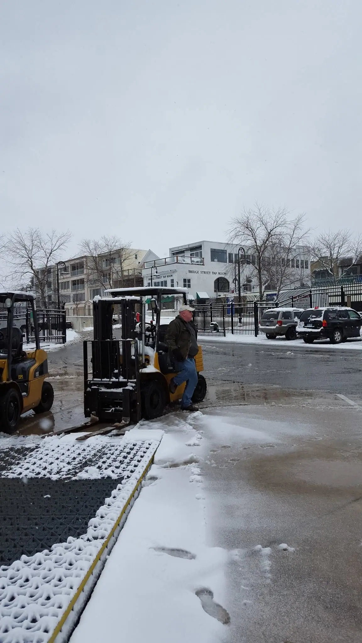 A worker operates a yellow forklift in a snowy paved lot beside a fenced commercial building.