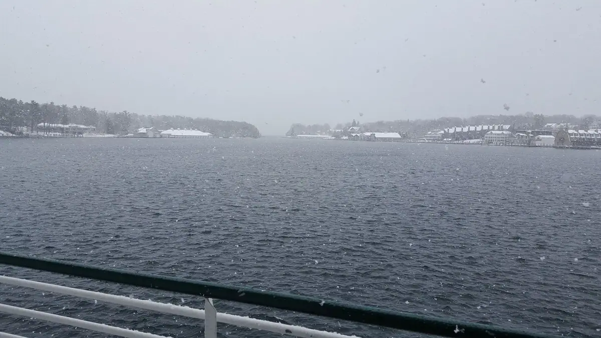 Snow falls over open water toward a tree-lined shore with clustered buildings, viewed past a vessel railing.