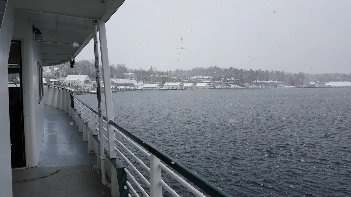 Snow falls across a waterfront viewed from a ferry deck, with houses and trees along the shore.