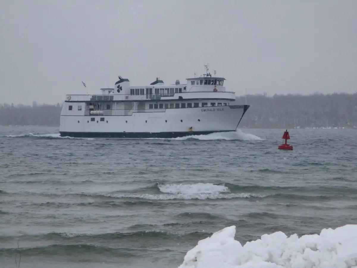 White passenger ferry underway beside a red navigation buoy, with snowy shore and rough water in the background.