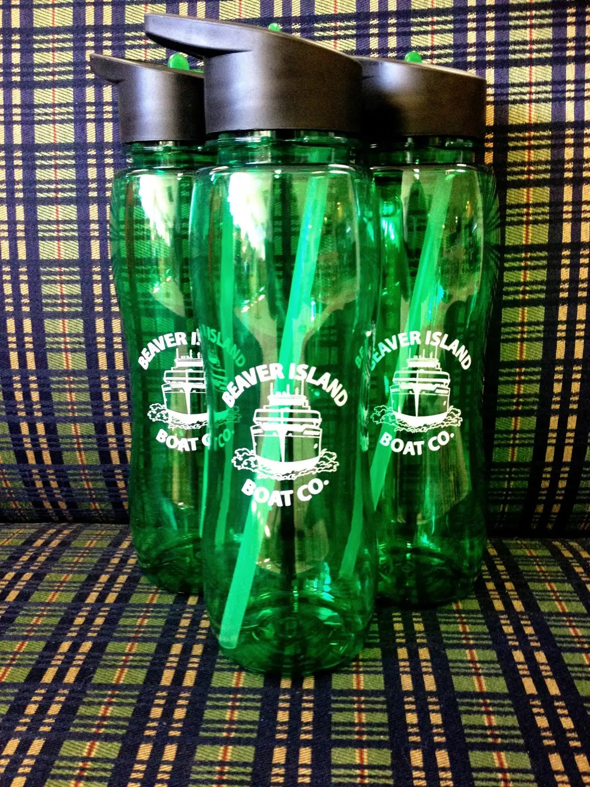 Three green reusable bottles with black flip-top lids and white boat logos rest on plaid fabric.