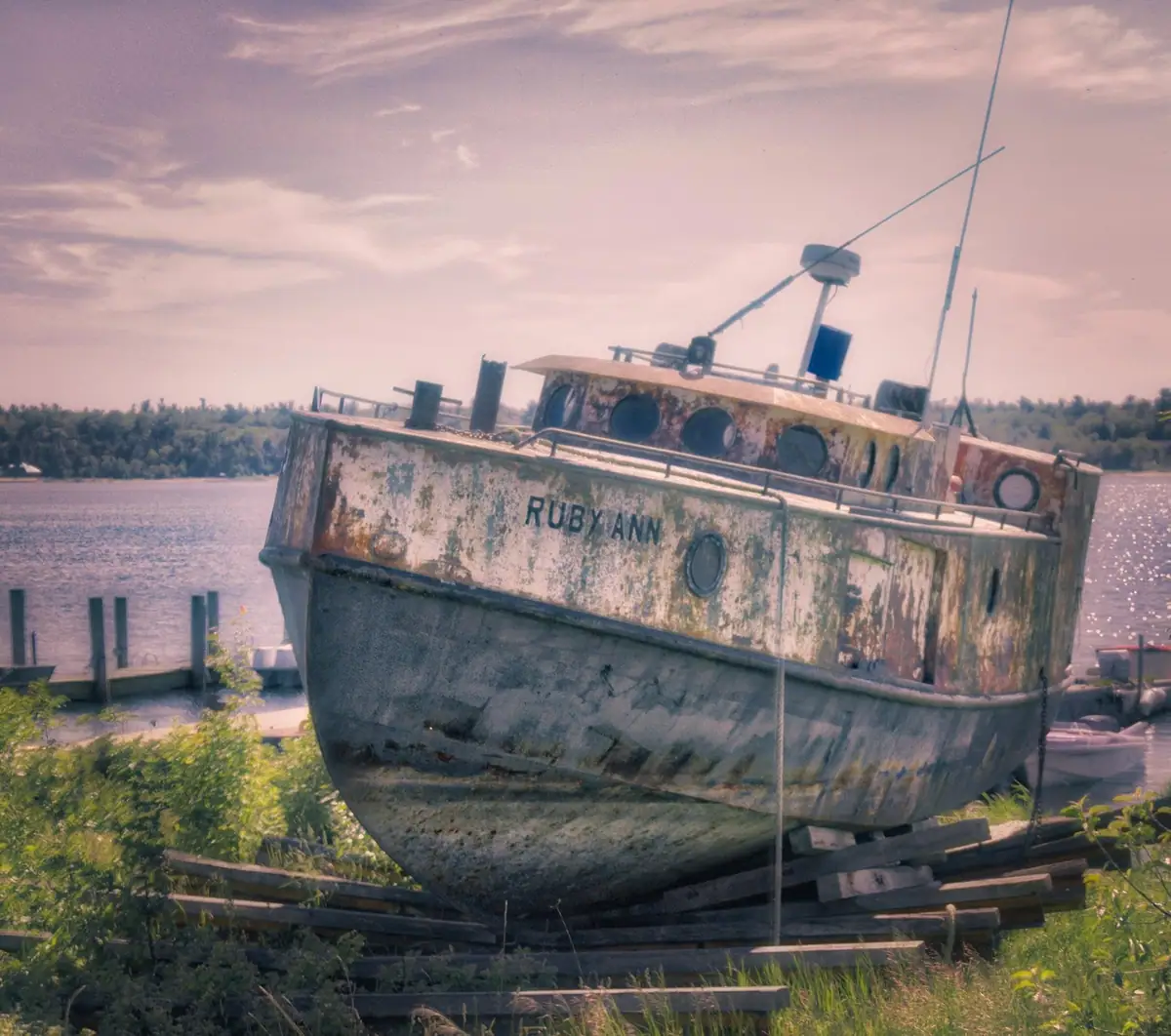 A rusted, weathered boat marked “RUBY ANN” rests on wooden supports beside the water.