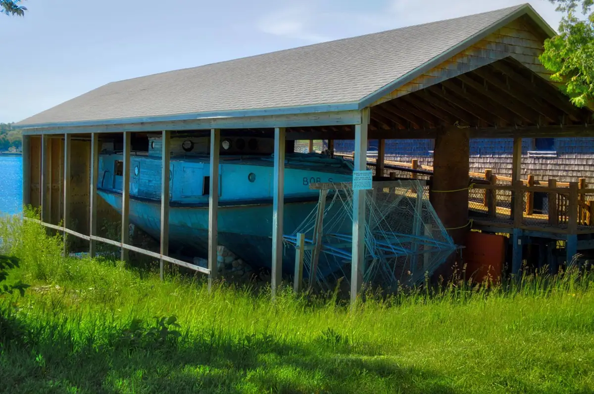 A blue boat rests beneath a wooden shelter beside the water, with fishing gear and a small sign in front.