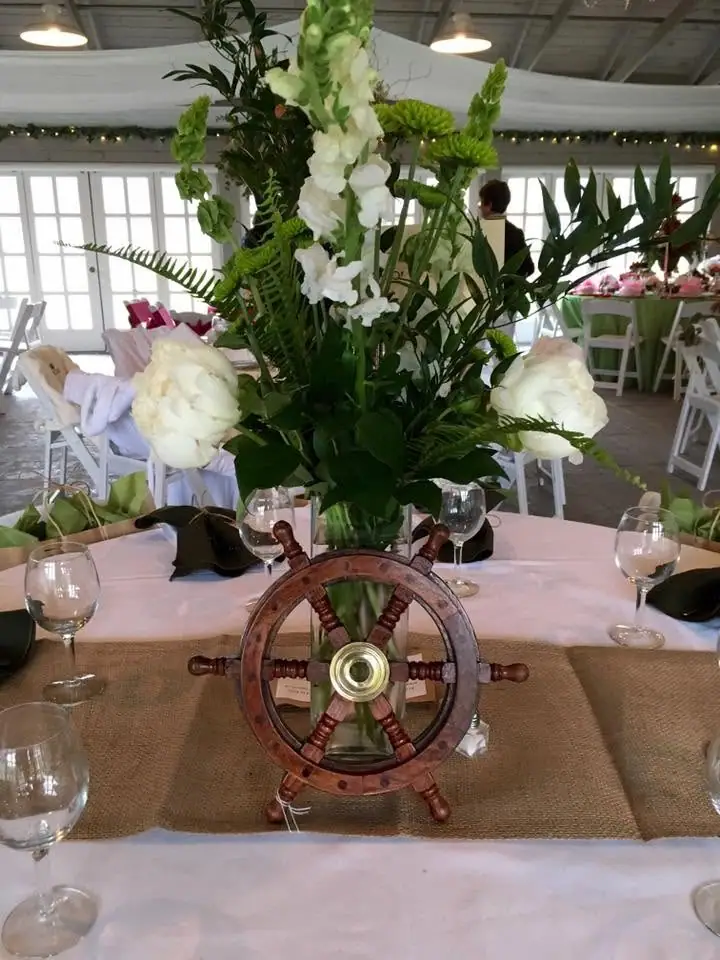 A decorated banquet table has a tall floral centerpiece and a wooden ship's wheel ornament.