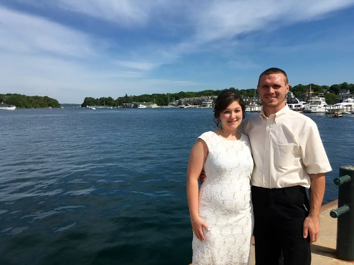A couple in formal attire stands on a pier beside a harbor filled with boats.