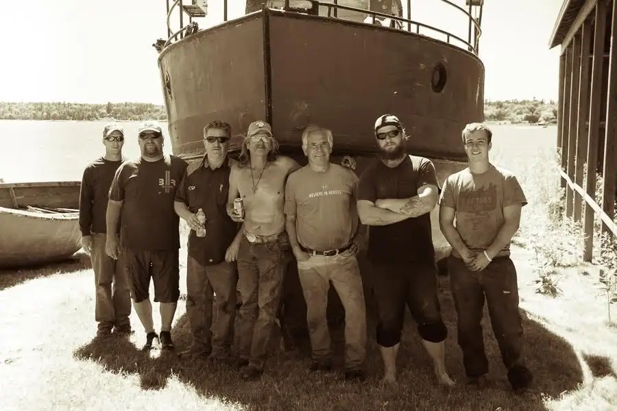 Seven men pose in front of a large vessel beside a small boat, water, wooded shoreline, and a wooden structure.