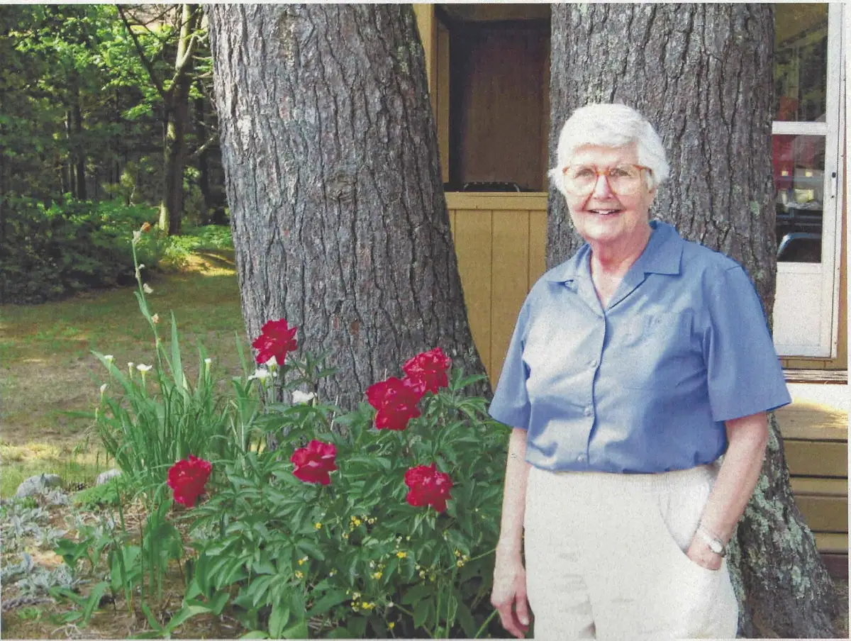 Older woman standing beside red flowers and large trees outside a wood-sided building.