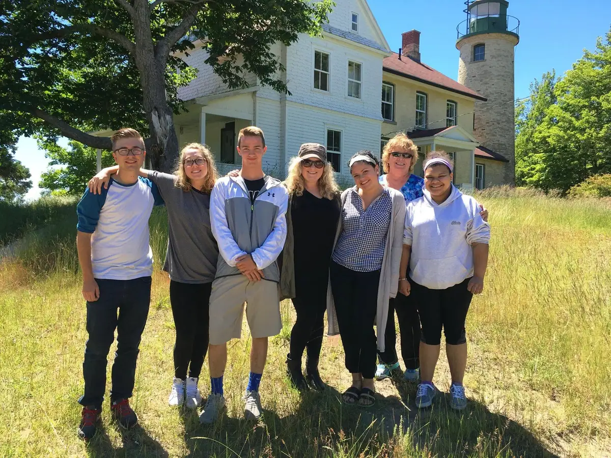 Seven people stand in tall grass beside a white house and stone lighthouse tower.