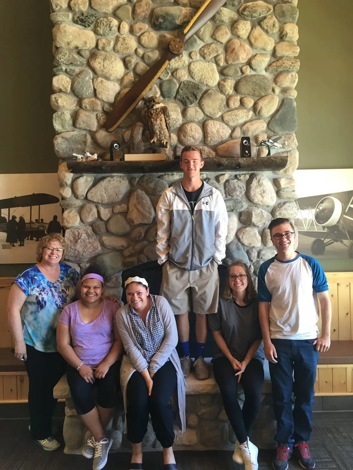Six people pose in front of a stone fireplace decorated with an owl, aircraft models, and a mounted propeller.