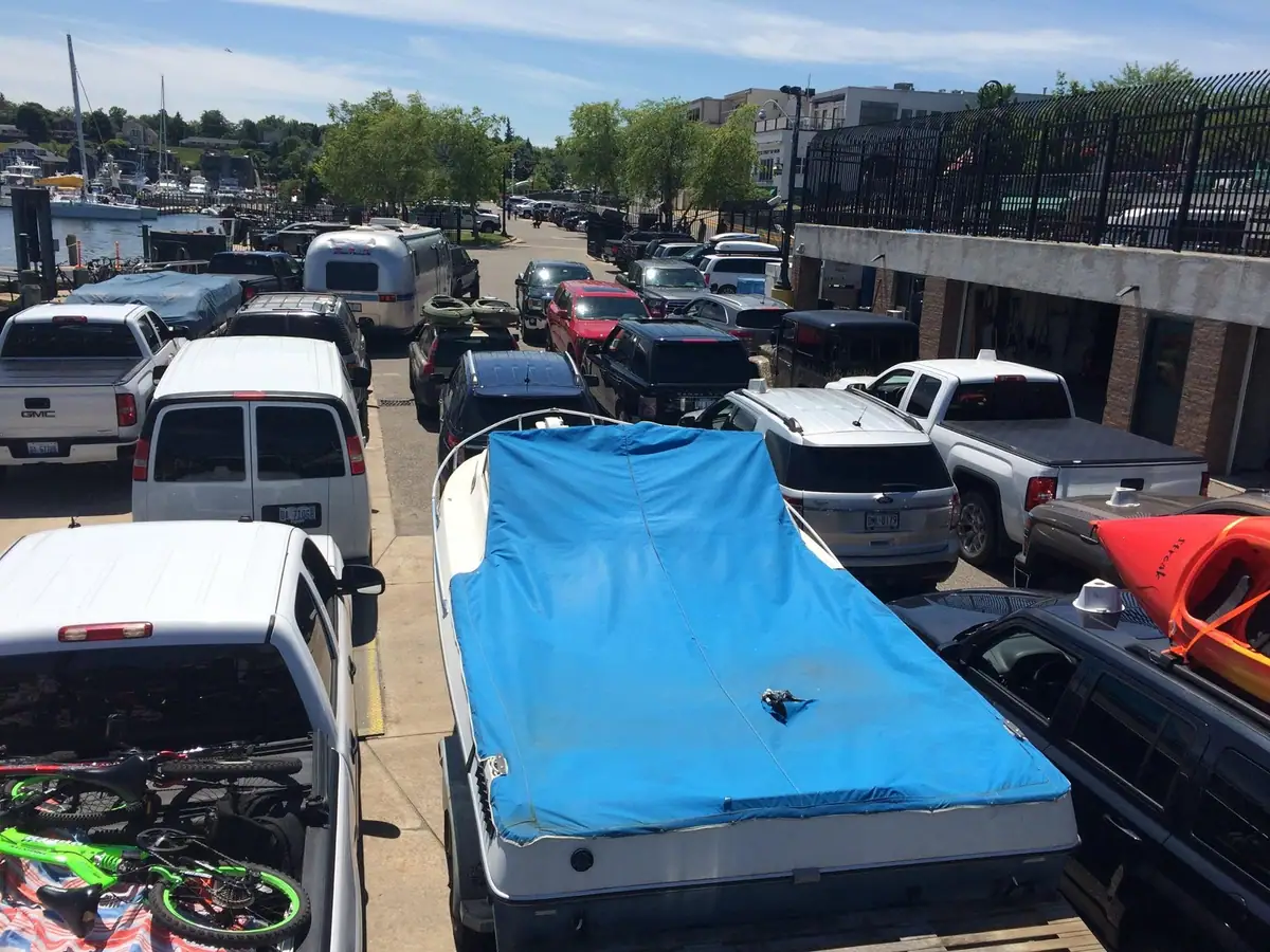 A crowded waterfront parking area with trucks, cars, bicycles, boats, and a blue-covered trailer.