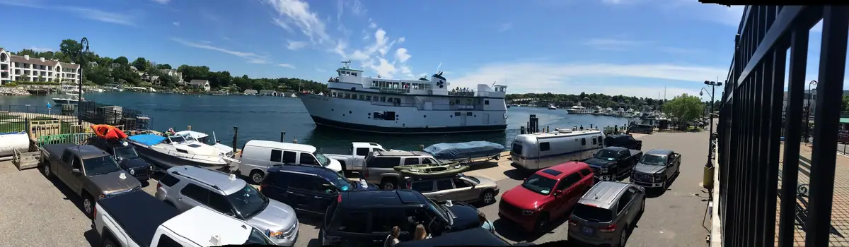 A white passenger ferry passes a waterfront crowded with parked vehicles, trailers, and small boats.