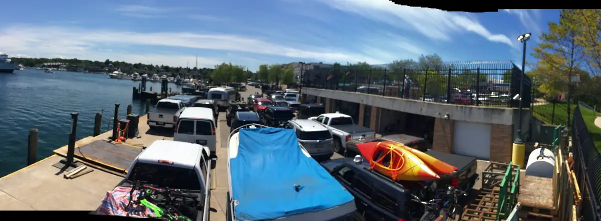 A waterfront parking area beside a dock, with pickup trucks, kayaks, and a blue-covered boat.