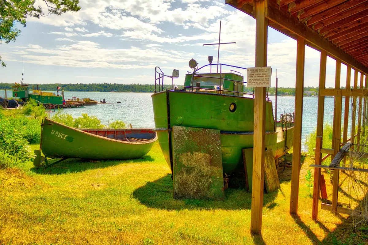 Green boats rest on grass beside a roofed wooden structure, with open water and a wooded shoreline beyond.