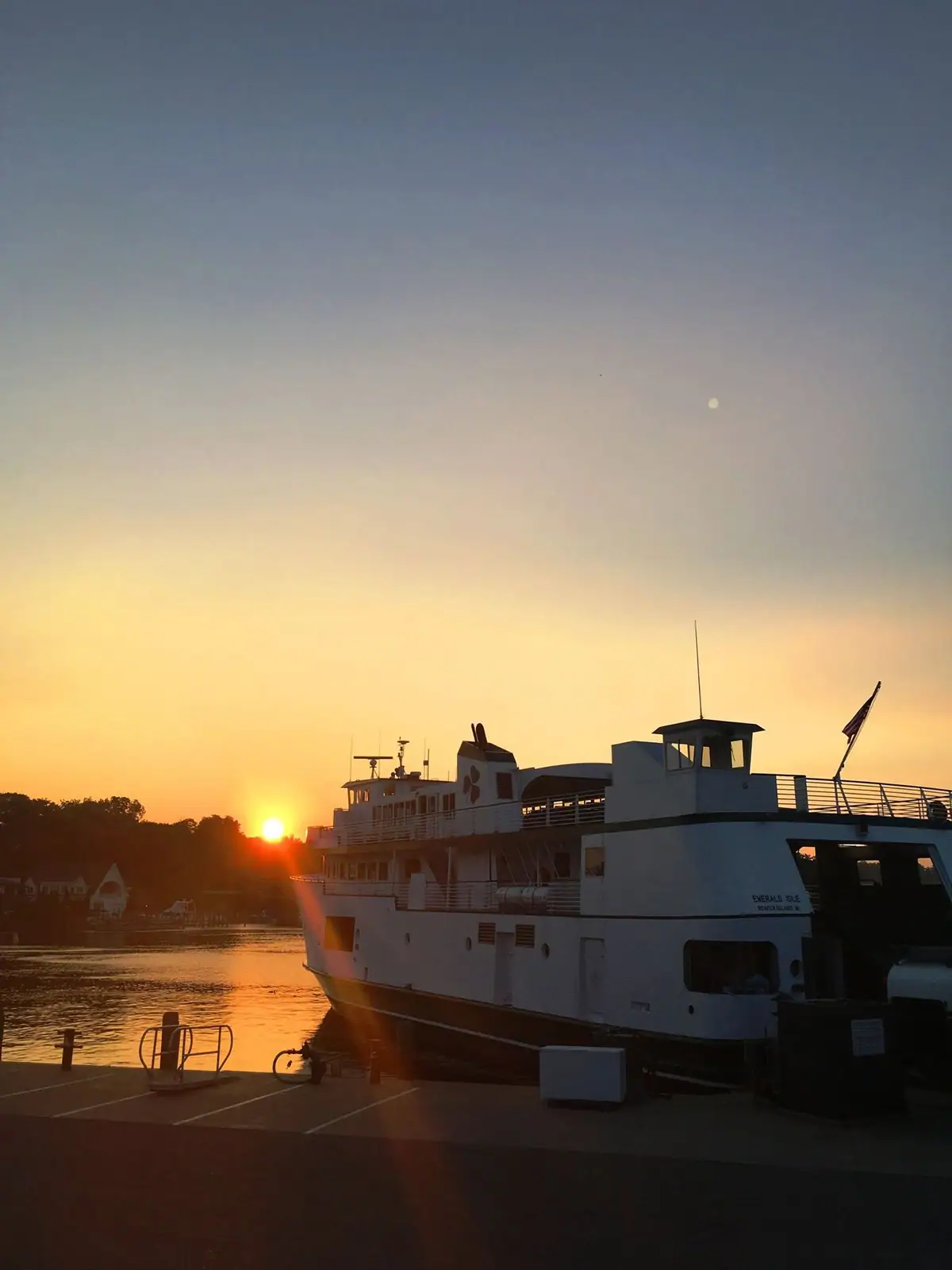 A white passenger ferry is docked beside a pier as the sun rises behind a tree-lined shore.
