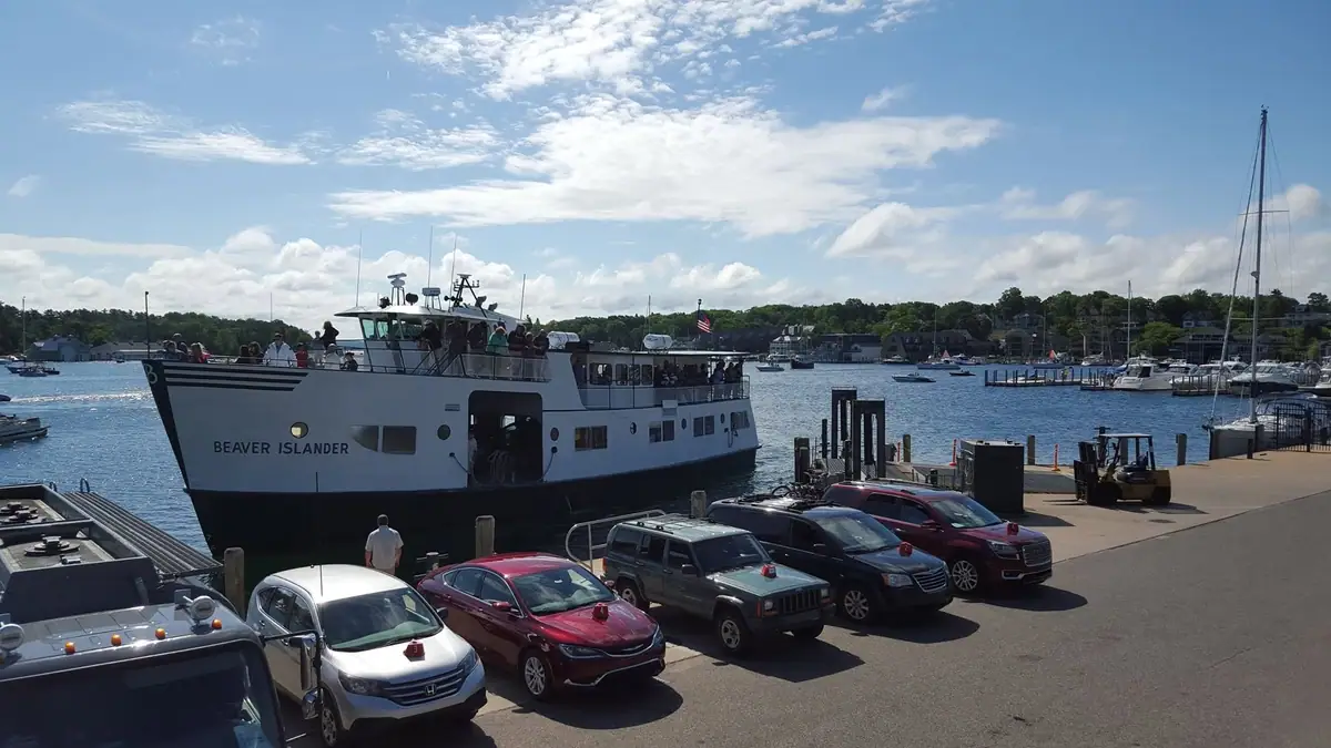 A passenger ferry is docked beside a pier with cars, a forklift, boats, and people visible in a sunny harbor.