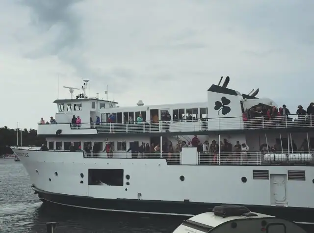 A crowded white passenger ferry with two decks moves across gray water beneath an overcast sky.