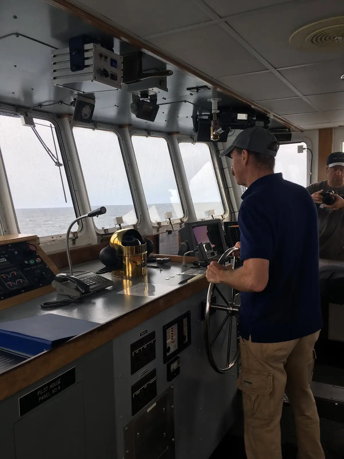 Two people are visible in a vessel's pilot house, where one stands at the wheel beside navigation equipment.