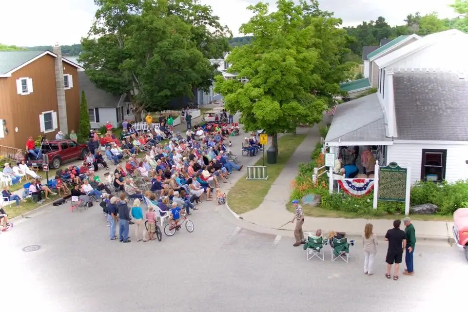 An elevated view shows a large crowd seated in lawn chairs along a residential street beside a decorated white building.